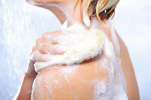 A girl washes herself in the shower with a soapy sponge.
